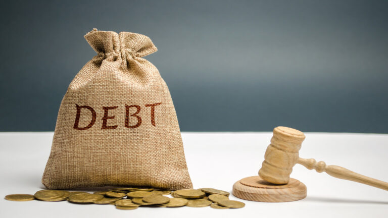 A burlap sack labeled "DEBT," a wooden gavel, and scattered coins on a gray surface with a blue background.