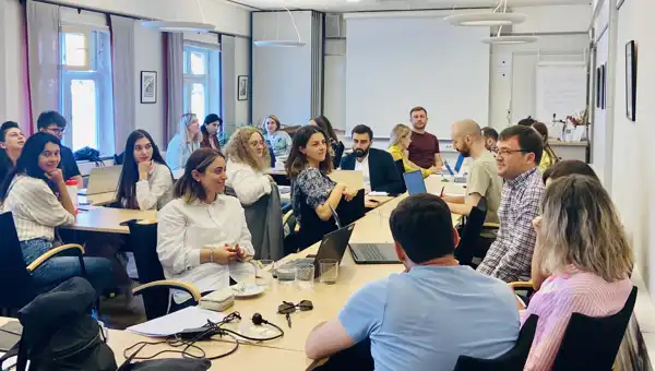 : Image showing a diverse group of young professionals in a collaborative setting, possibly with Swedish and Eastern European flags or digital charts, symbolizing international training and leadership development.